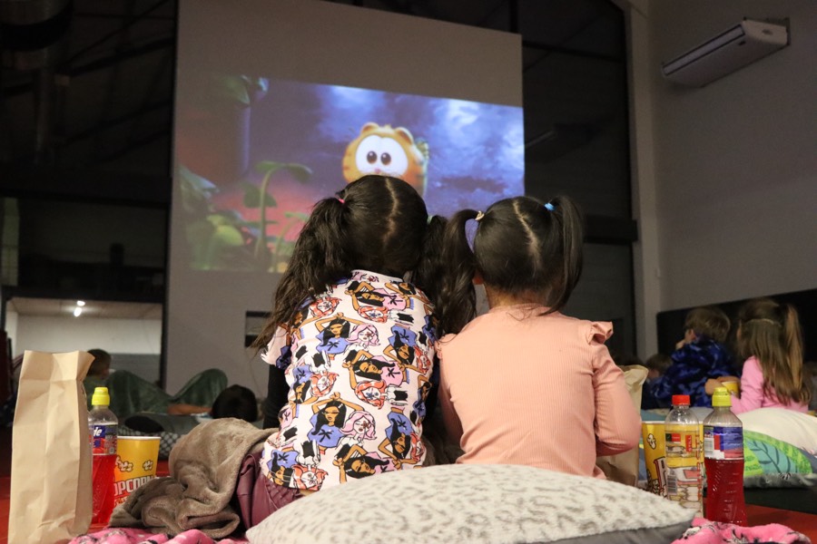 Two children in pajamas watching a movie on the big screen, wrapped in blankets