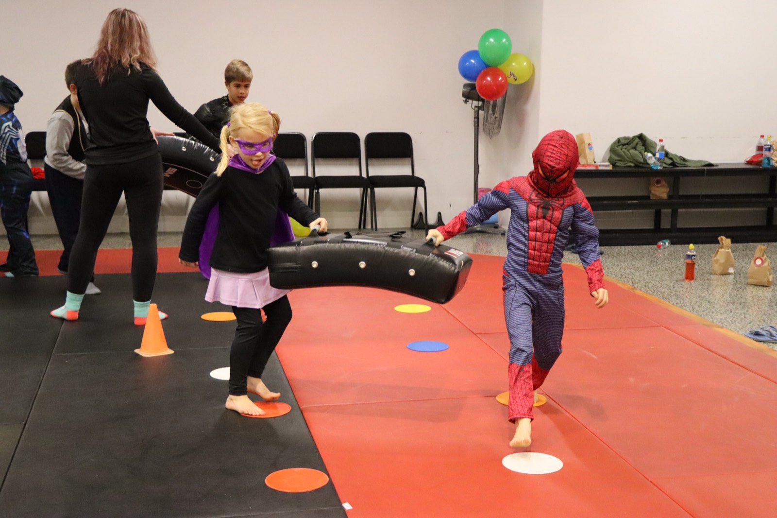 A child in a superhero costume kicking a training pad held by another child in a cape and mask, on the mats.